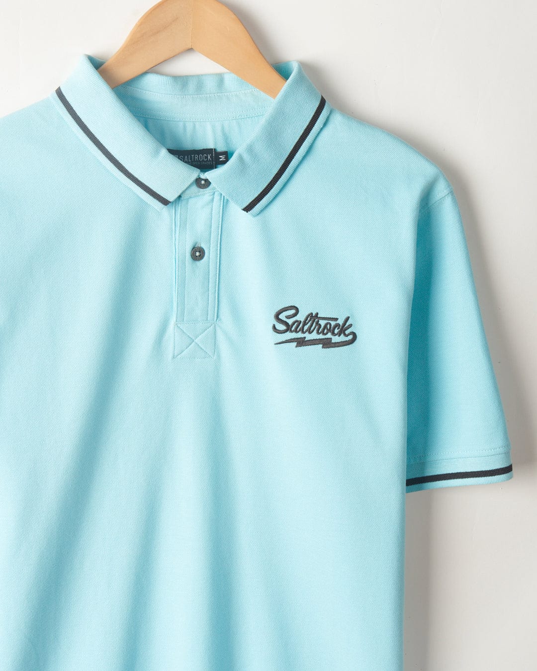 A light blue Saltrock Strike Logo - Mens Short Sleeve Polo Shirt with black stripes on the collar and sleeve edges. It has a "Saltrock" logo embroidered on the left chest. The polo shirt is hung on a wooden hanger against a white wall.