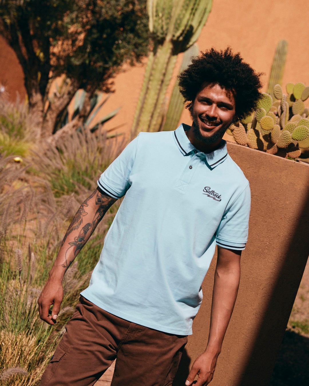 A man with curly hair and tattoos on his right arm wears a Saltrock Strike Logo - Mens Short Sleeve Polo Shirt - Light Blue and brown pants, standing outdoors near cacti and plants.