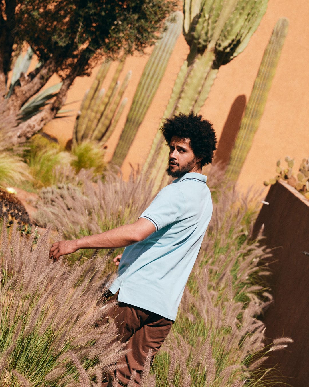 A person in a Strike Logo - Mens Short Sleeve Polo Shirt - Light Blue, featuring the Saltrock embroidered logo, stands among tall grasses, with cacti and an orange wall in the background.