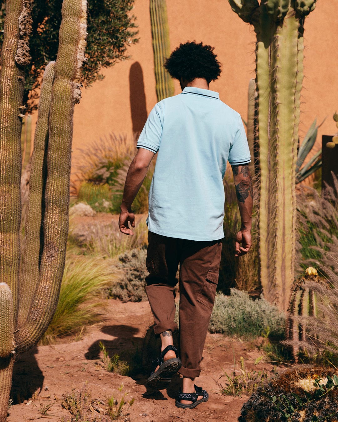 A person with an afro hairstyle, wearing a 100% cotton Saltrock Strike Logo - Mens Short Sleeve Polo Shirt - Light Blue and brown pants, walking away through a desert garden filled with tall cacti and other desert plants.