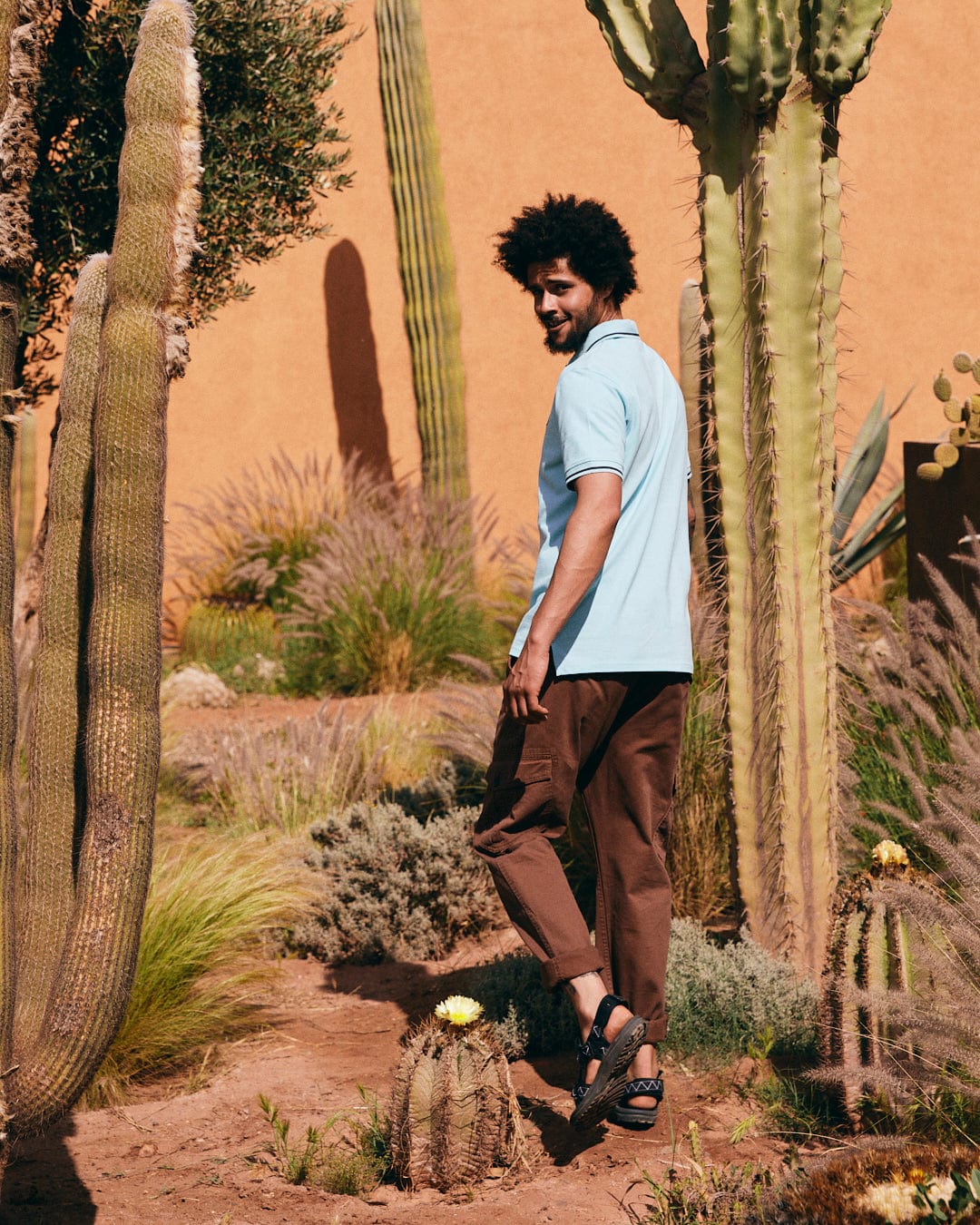 A person wearing a Strike Logo - Mens Short Sleeve Polo Shirt - Light Blue by Saltrock and brown pants walks through a garden with various cacti and plants, smiling and glancing over their shoulder.