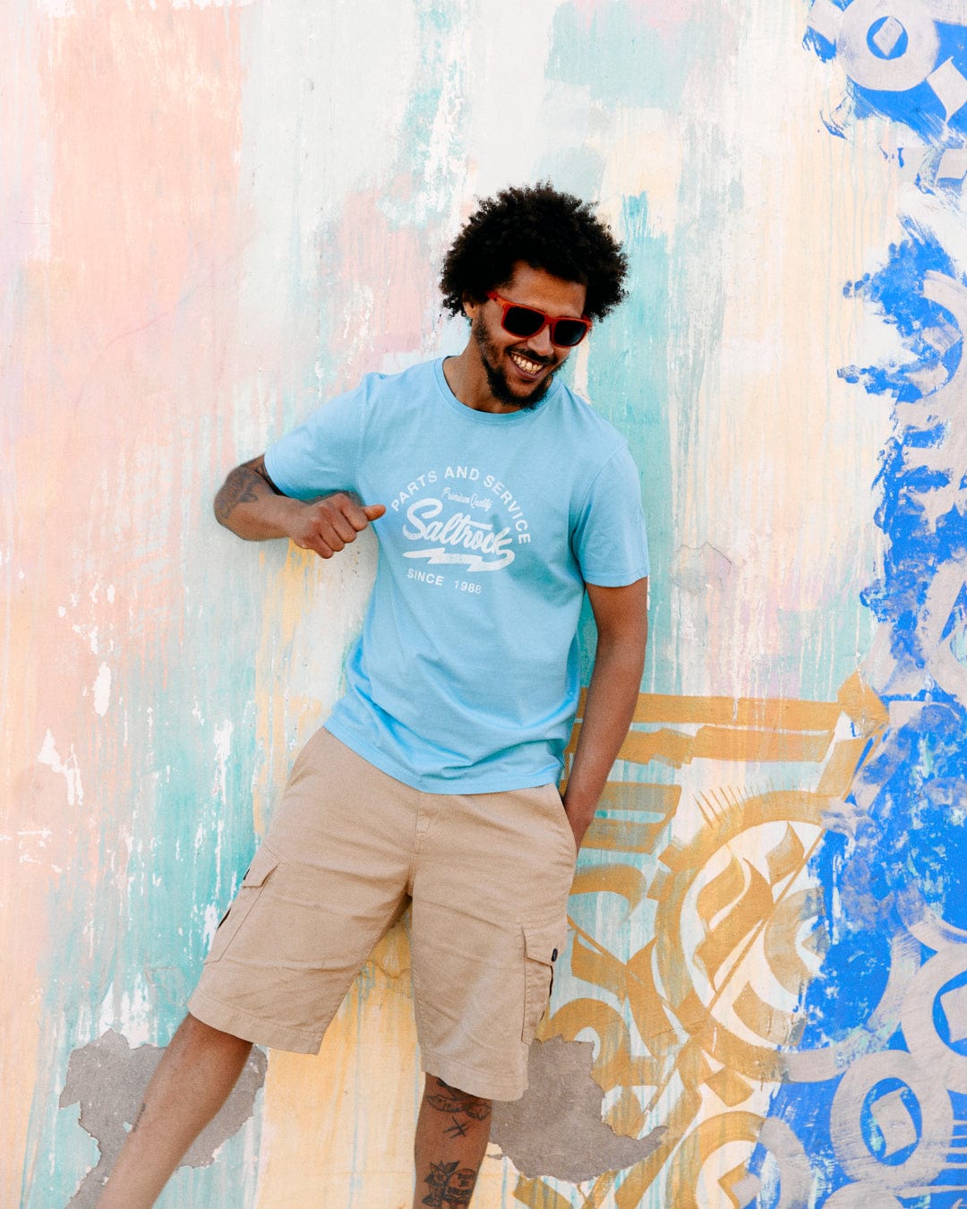 A joyful man in sunglasses and a Last Stop Motel - Mens T-Shirt in Light Blue from Saltrock laughs while leaning against a colorful mural wall.
