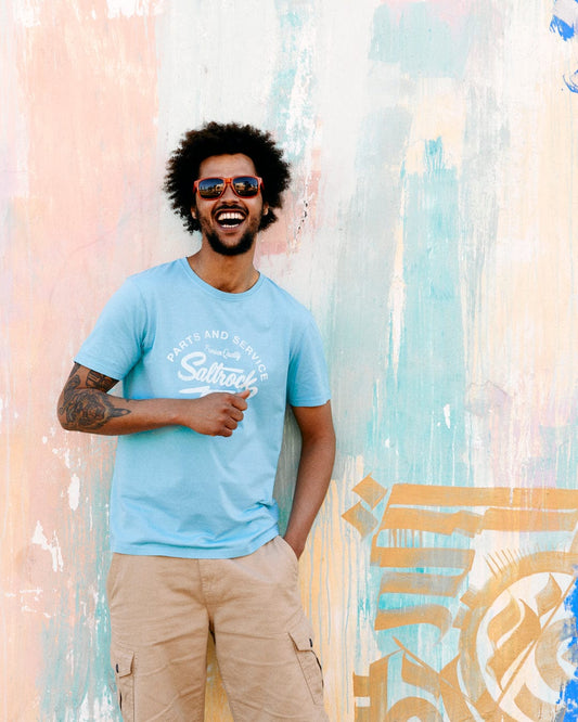 A joyful man with an afro wearing sunglasses and a blue Saltrock 100% Cotton Last Stop Motel t-shirt stands against a colorful mural.