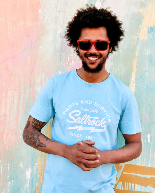 A man with curly hair wearing sunglasses and a Saltrock Last Stop Motel - Mens T-Shirt - Light Blue, smiling in front of a colorful wall.