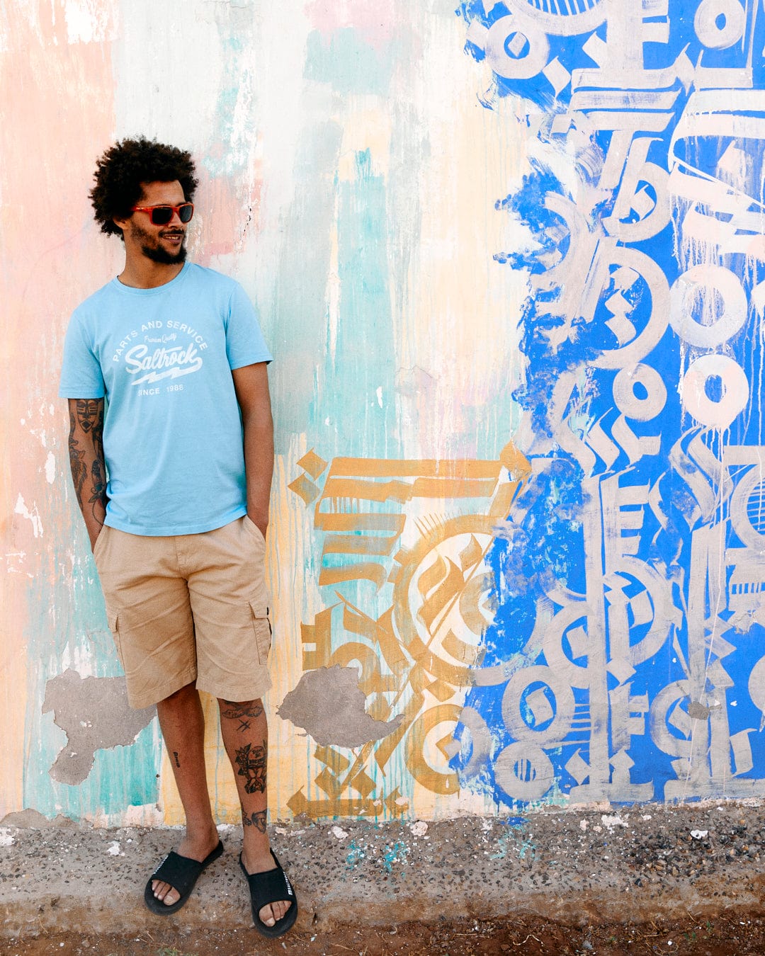 A man in a Last Stop Motel - Mens T-Shirt - Light Blue from Saltrock and beige shorts stands smiling against a colorful graffiti wall.