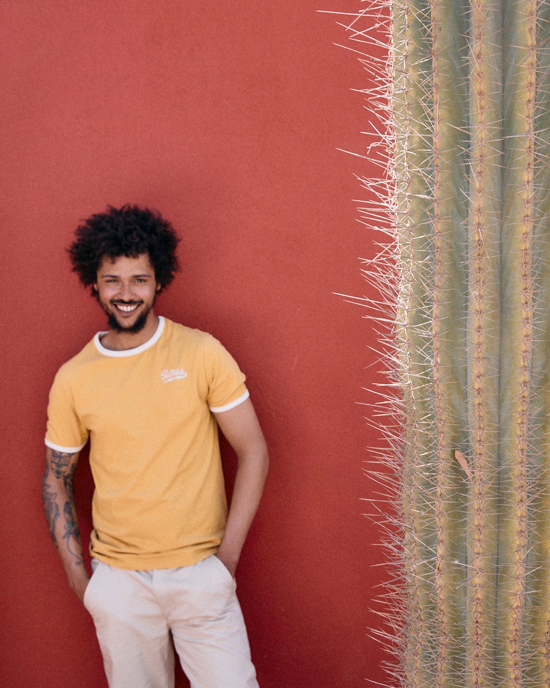 A person with curly hair and a beard, wearing a yellow Strike Ringer - Mens Short Sleeve T-Shirt by Saltrock and light pants, stands smiling against a red wall next to a large cactus.