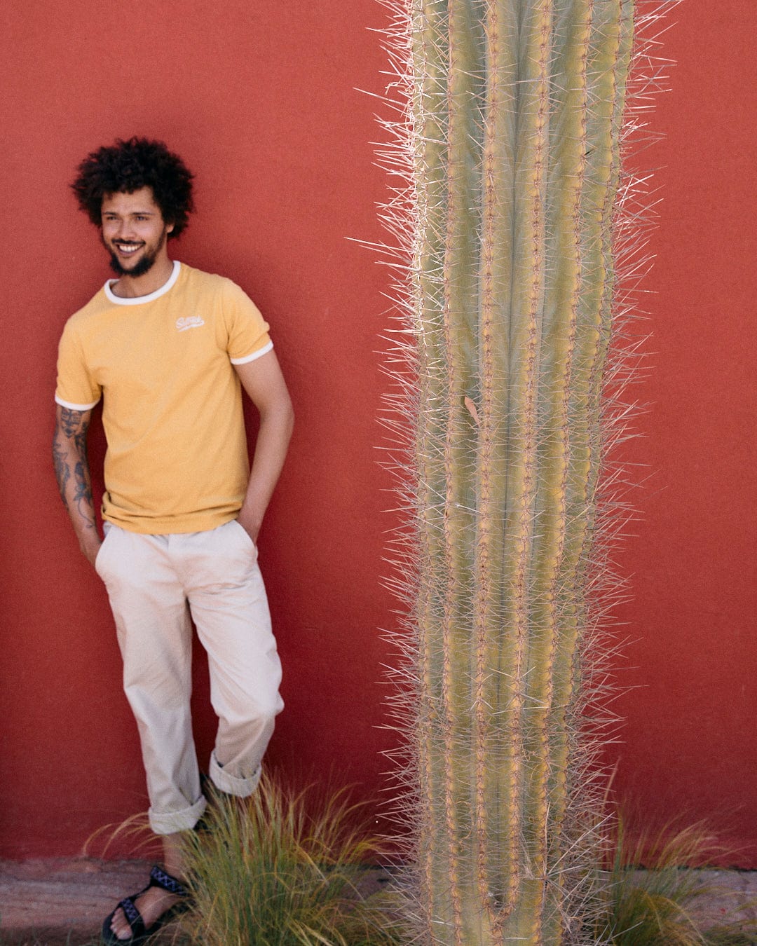 A person stands smiling against a red wall next to a tall cactus. They have curly hair, beige pants, and are wearing sandals. They are also wearing a yellow shirt with Saltrock embroidery called the Strike Ringer - Mens Short Sleeve T-Shirt by Saltrock.