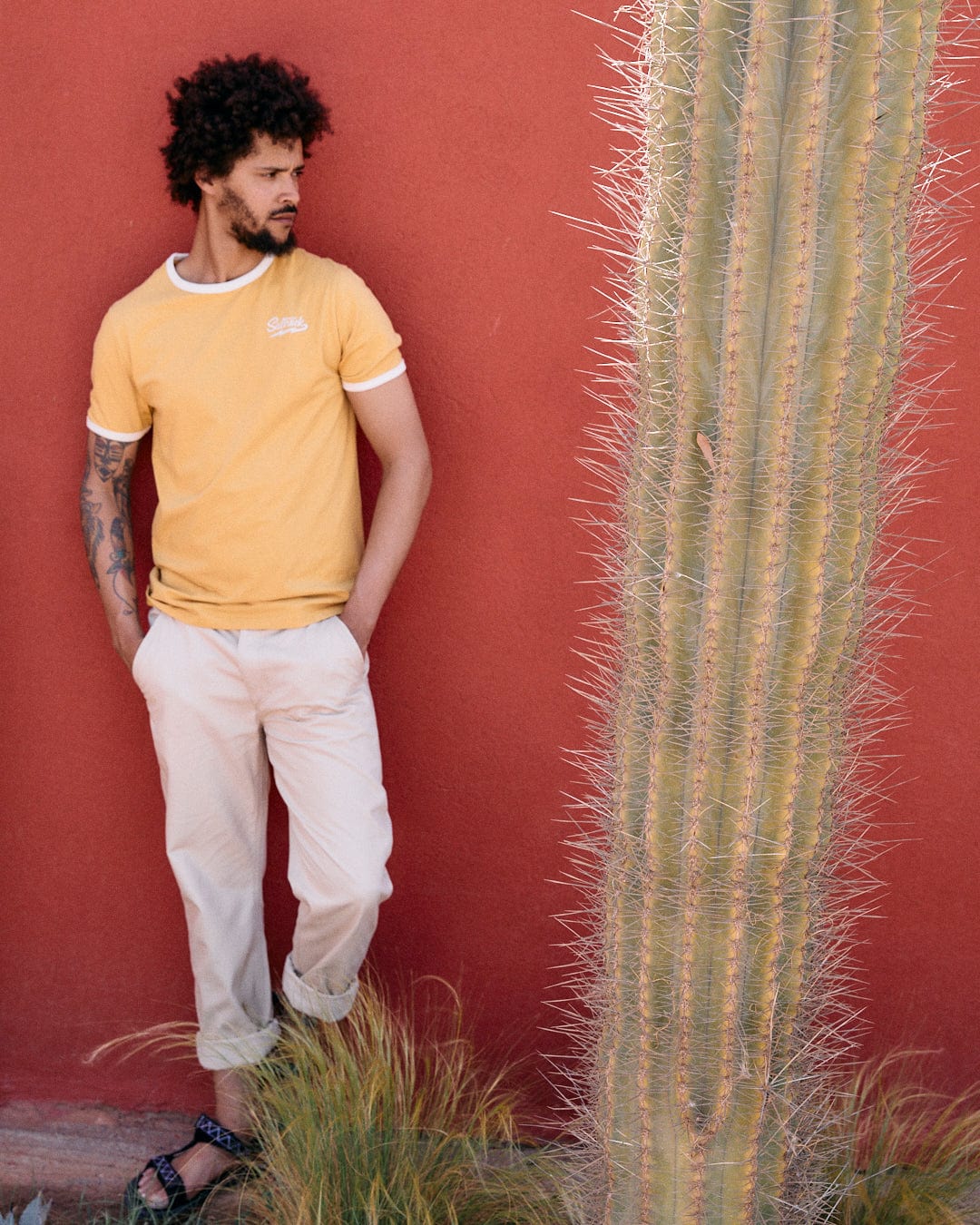 A man in a Strike Ringer - Mens Short Sleeve T-Shirt - Yellow by Saltrock and beige pants stands next to a tall cactus, leaning against a red wall.