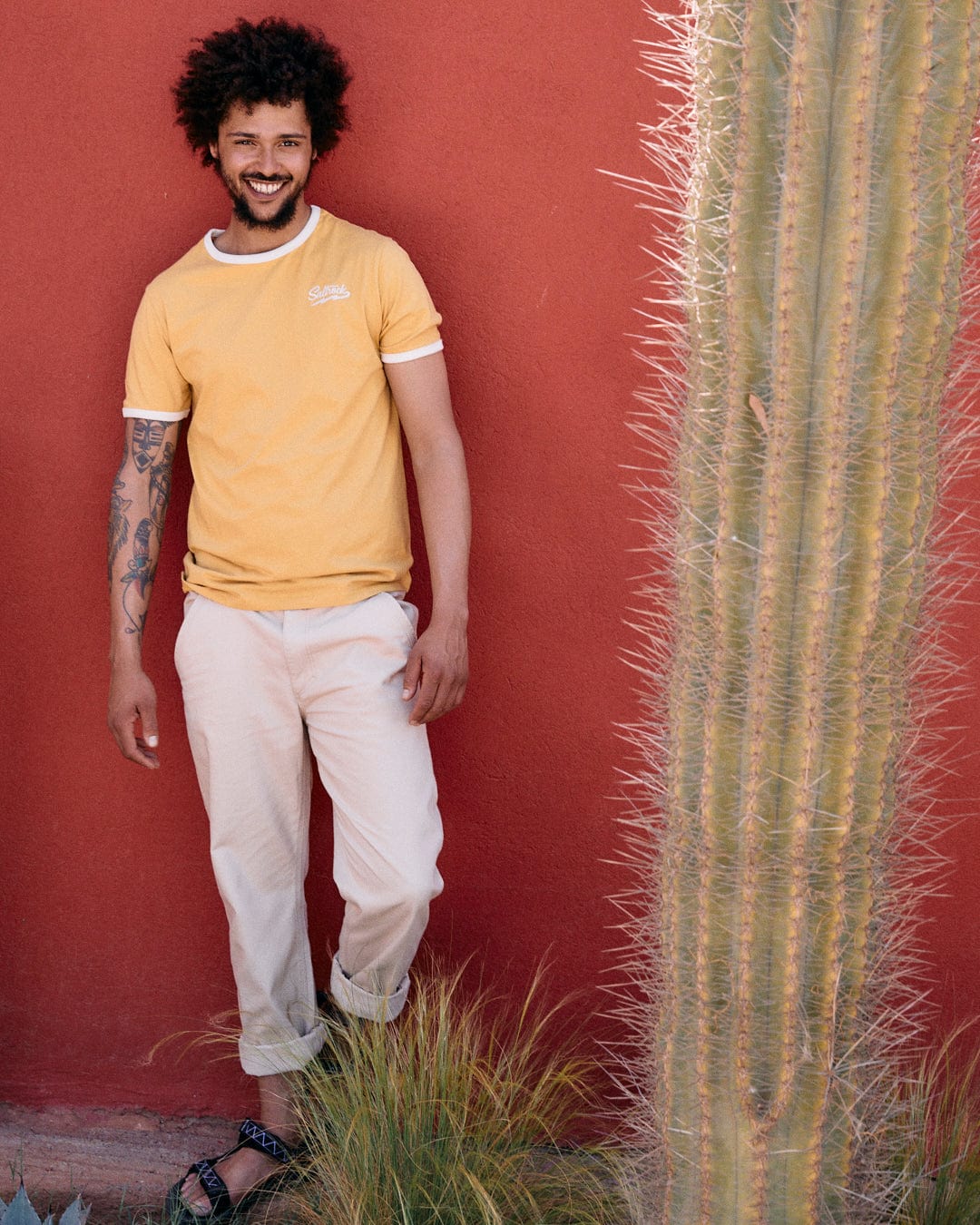 A man in a Strike Ringer - Mens Short Sleeve T-Shirt - Yellow from Saltrock and beige pants stands smiling next to a cactus against a red wall.