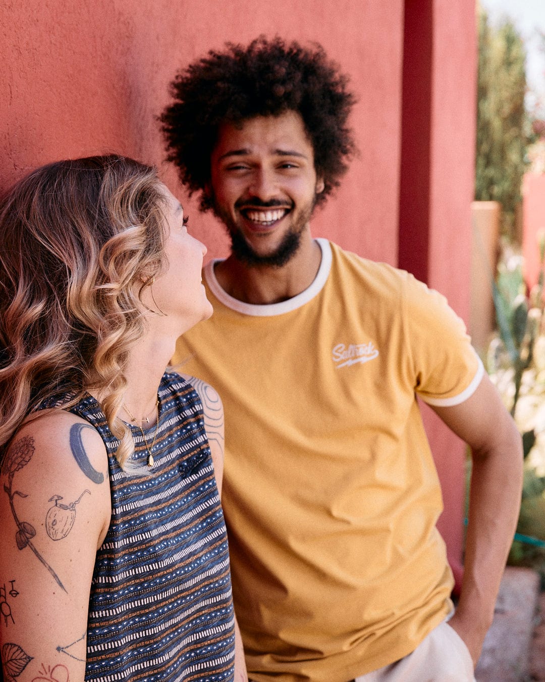 A woman with tattoos and a man in a yellow Strike Ringer - Mens Short Sleeve T-Shirt - Yellow from Saltrock smile while leaning against a red wall.