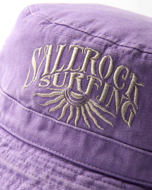 Close-up of embroidered lettering that reads "surfing" with a Saltrock sun design on a Sunburst Bucket Hat - Purple fabric background.