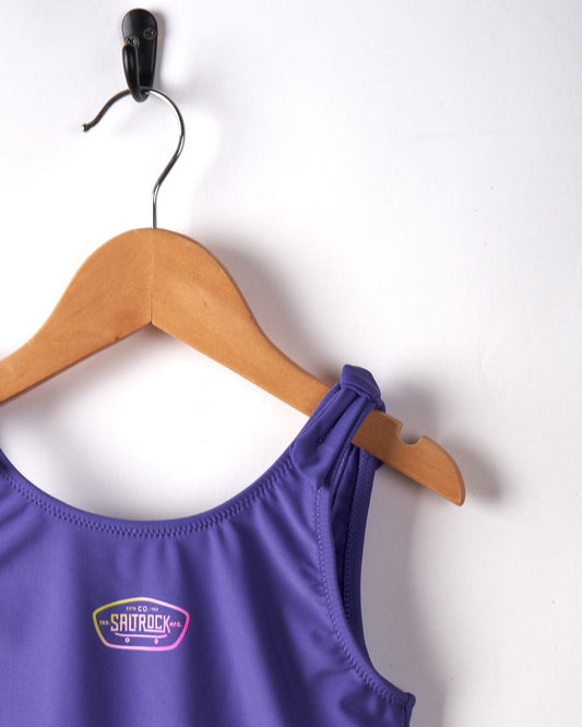 A purple Sunny Hardskate - Recycled Kids Swimsuit with a Saltrock gradient logo, hanging on a wooden hanger against a white background.
