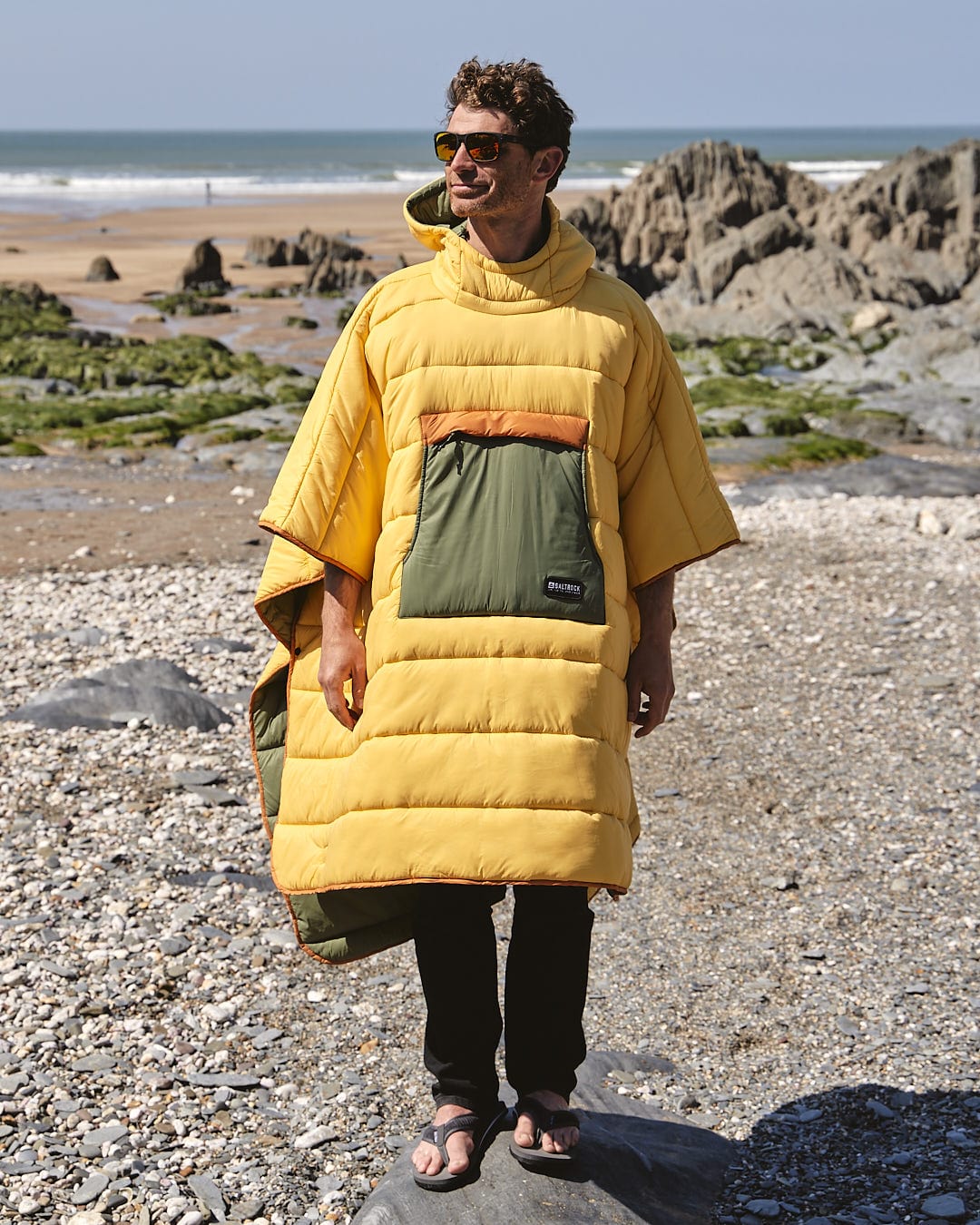 A man in a Sunset - Packable Reversible Poncho - Yellow by Saltrock standing on the beach.