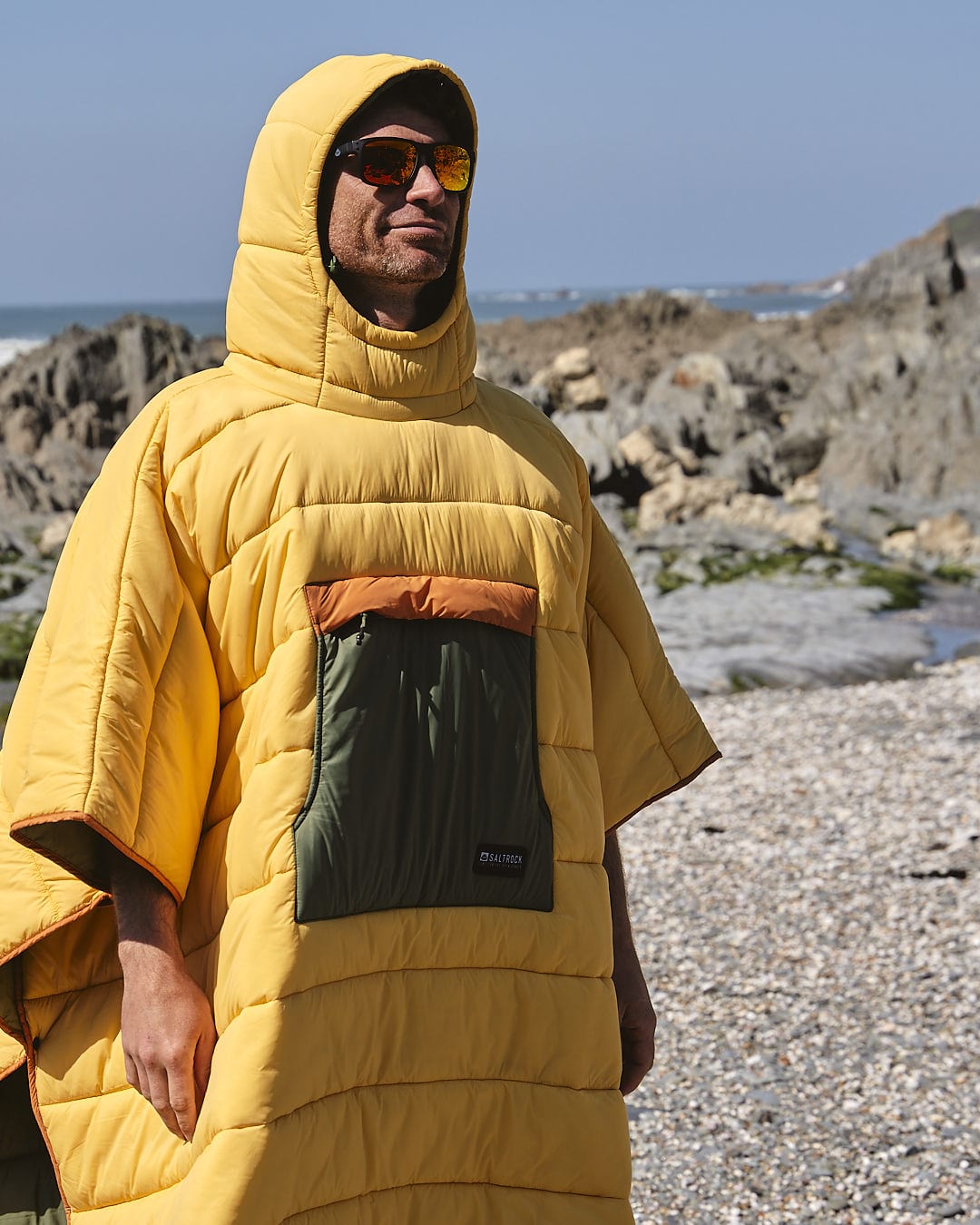 A man in a Saltrock Sunset - Packable Reversible Poncho - Yellow standing on the beach.