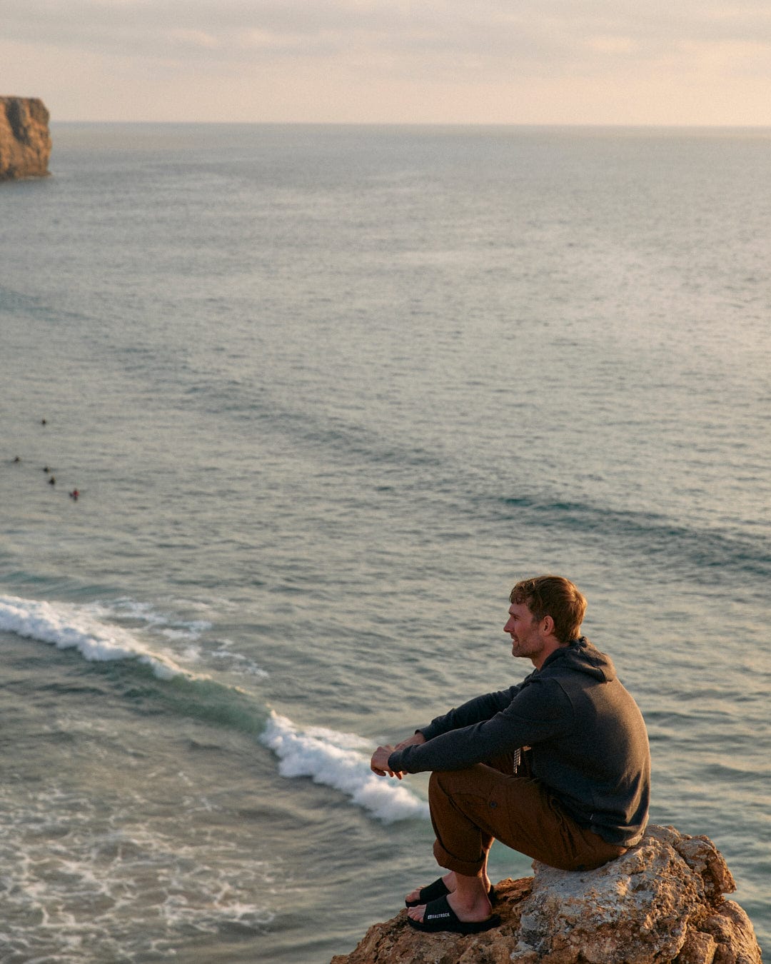 A man sitting on a cliff overlooking the ocean with a Saltrock Sun Sets - Mens Pop Hoodie - Charcoal.