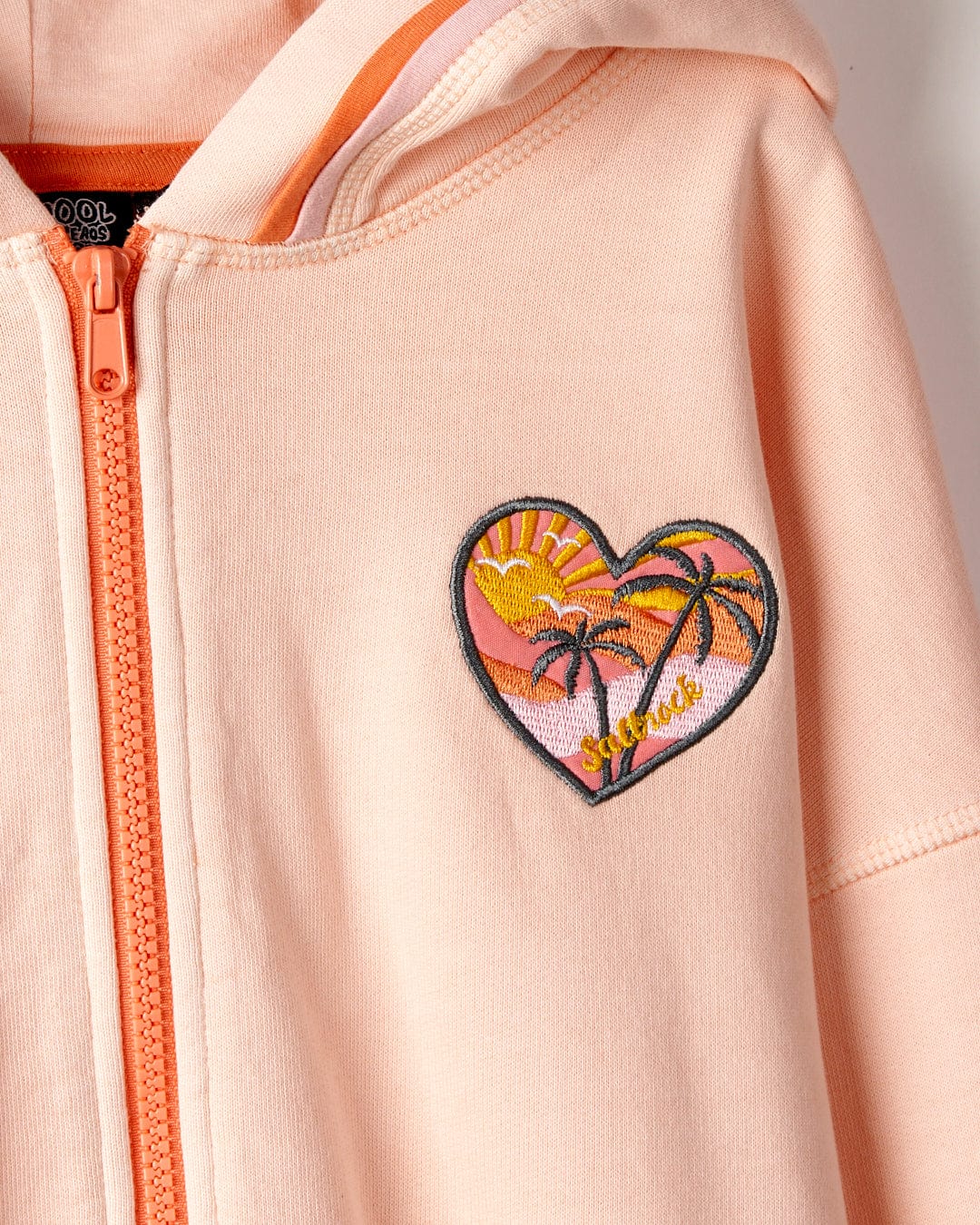 Close-up of a pink, 100% cotton Sunshine State - Kids Zip Hoodie in Peach by Saltrock with a heart-shaped patch featuring a tropical beach scene, complete with palm trees and sunset.