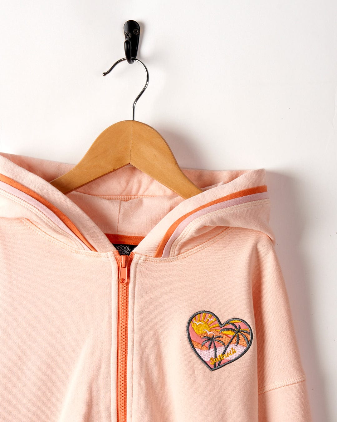 A Sunshine State - Kids Zip Hoodie in Peach with a tropical sunset patch over the left chest, hanging from a wooden hanger on a white background. Made from 100% cotton by Saltrock.