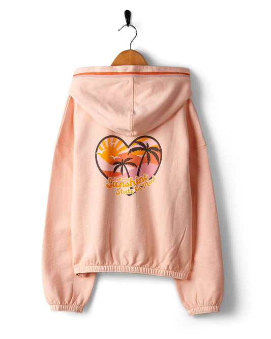A pink Sunshine State - Kids Zip Hoodie in Peach by Saltrock, featuring a "sunshine state of mind" graphic with palm trees and a sunset, displayed on a wooden hanger against a white background. This 100% cotton hoodie is machine wash.
