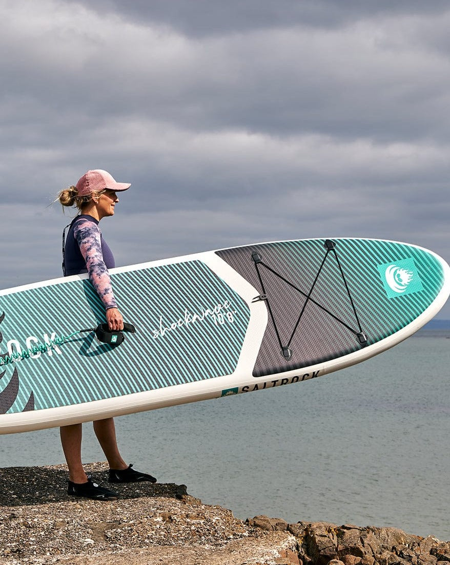 A woman carrying a Saltrock Shockwave - Inflatable Stand Up Paddle Board - Turquoise.