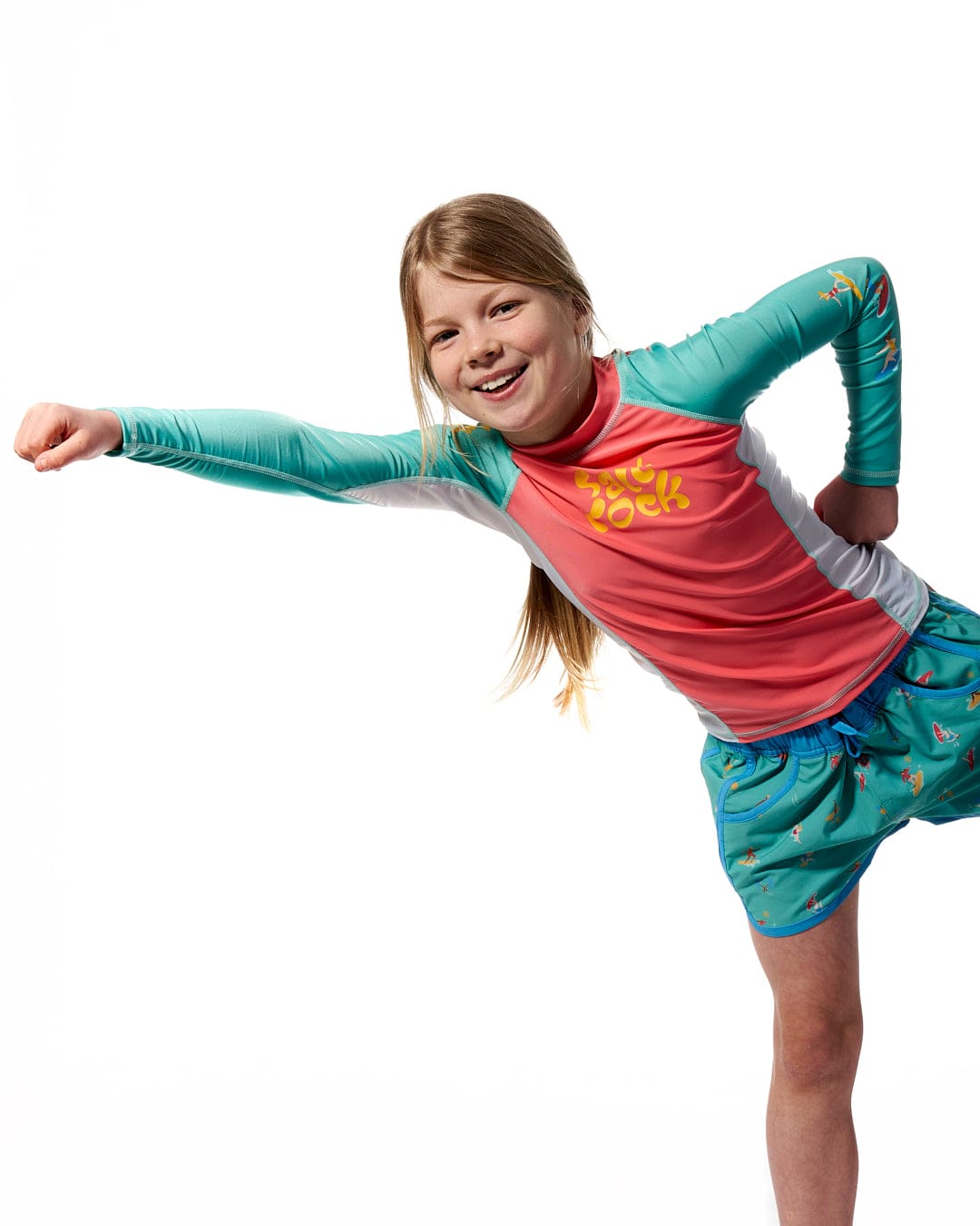 A young girl is posing for a photo wearing the Surf Sister - Kids Long Sleeve Rashvest in Coral/Turquoise by Saltrock.