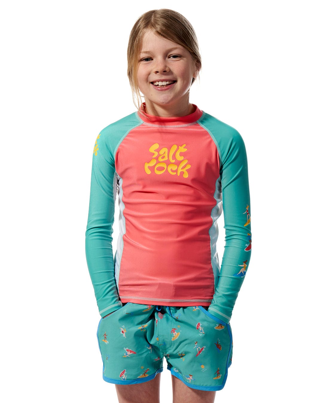 A young girl wearing a Surf Sister - Kids Long Sleeve Rashvest - Coral/Turquoise by Saltrock and shorts.