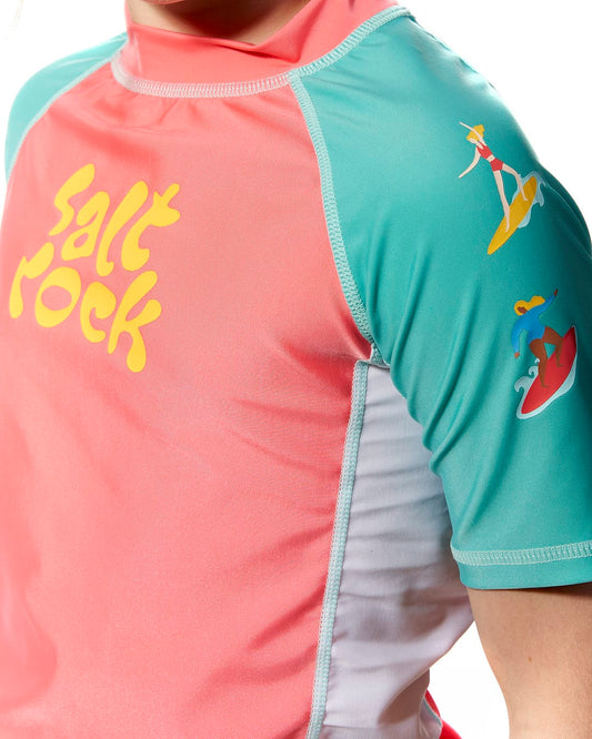 A young girl wearing a Saltrock Surf Sister - Kids Short Sleeve Rashvest in Coral/Turquoise.