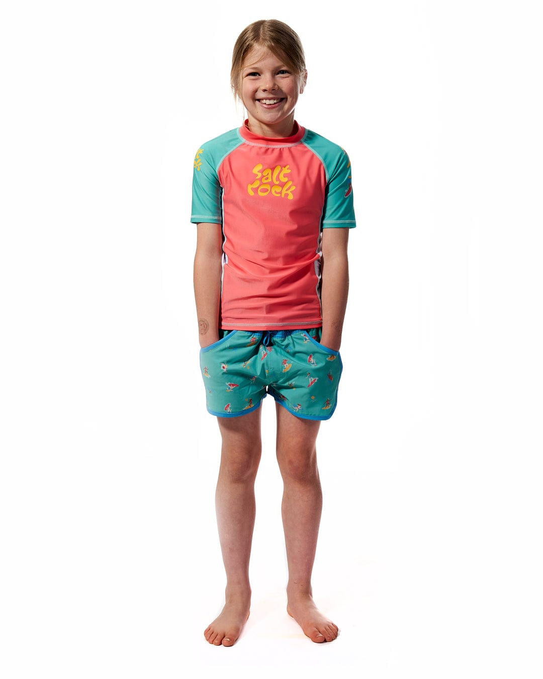 A young girl wearing a Saltrock Surf Sister - Kids Short Sleeve Rashvest in Coral/Turquoise and shorts.