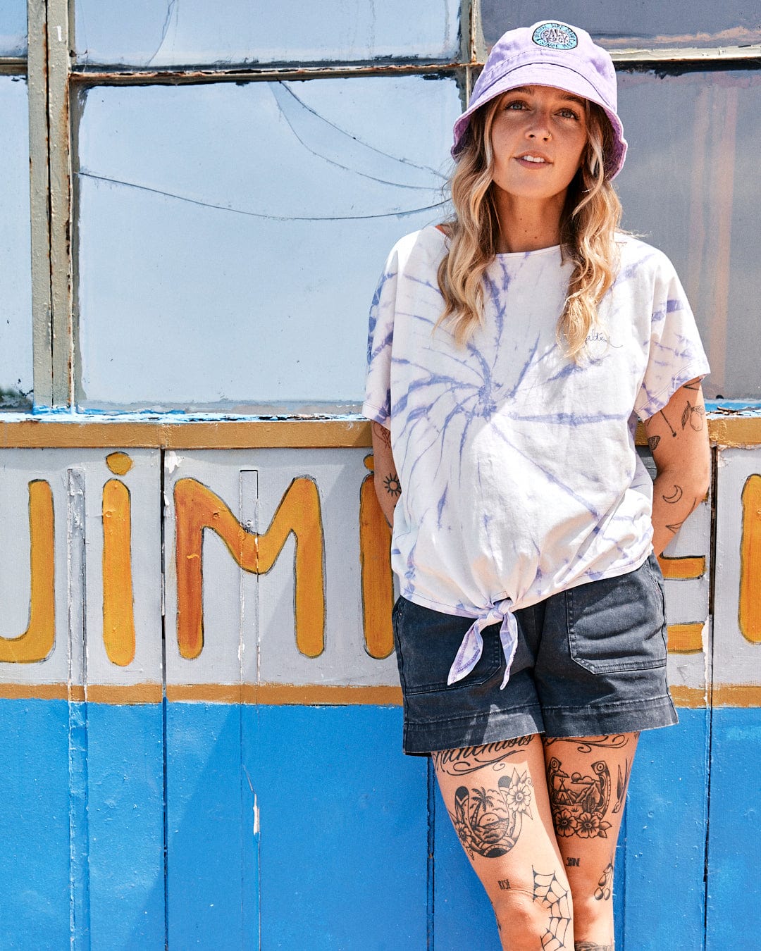 Woman in a Saltrock White/Purple Swirl - Womens Short Sleeve T-Shirt and denim shorts stands in front of a colorful vintage truck, wearing a purple hat, showcasing tattooed legs.