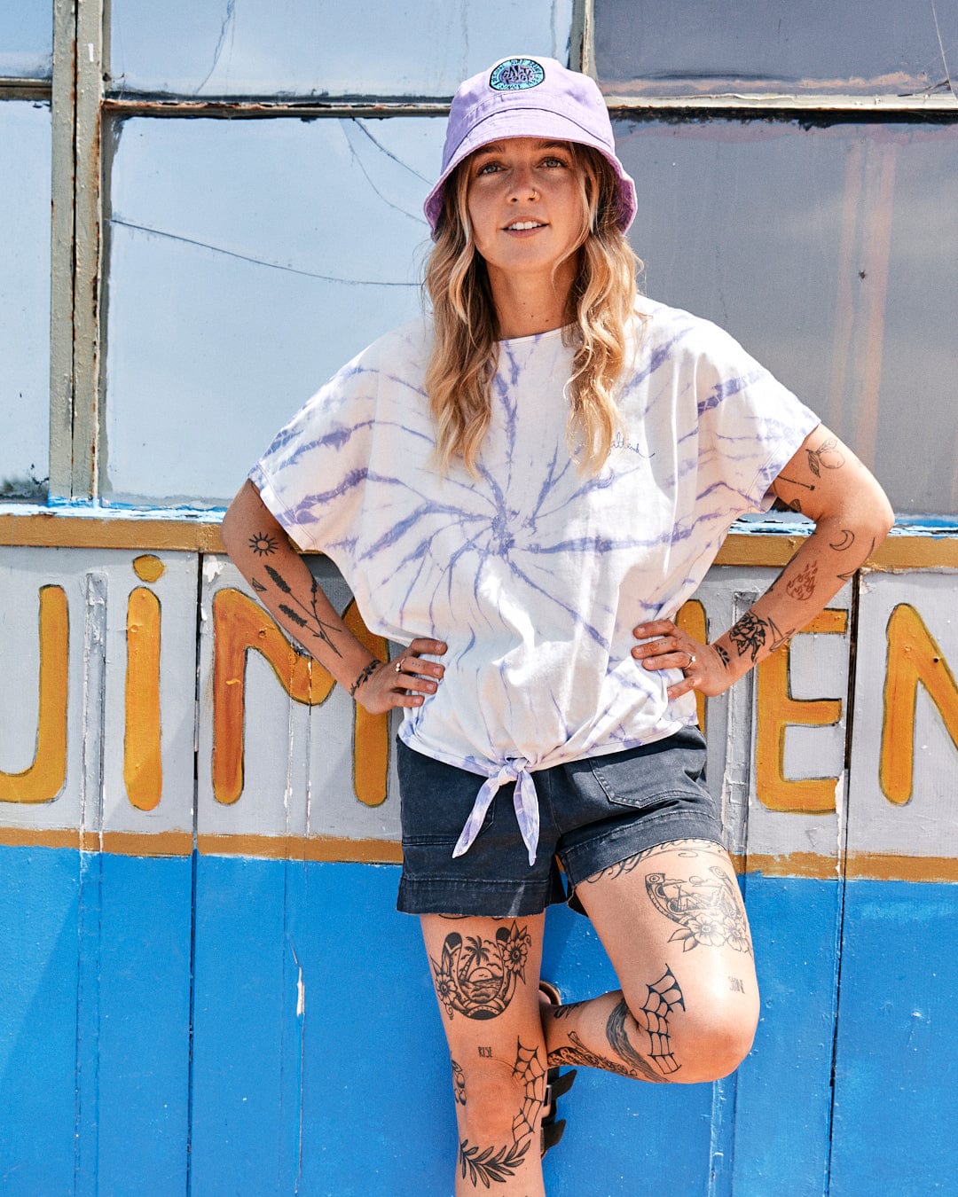 Woman in Saltrock Swirl - Womens Short Sleeve T-Shirt - Tie Dye White/Purple and denim shorts standing in front of a colorful, graffiti-covered wall.