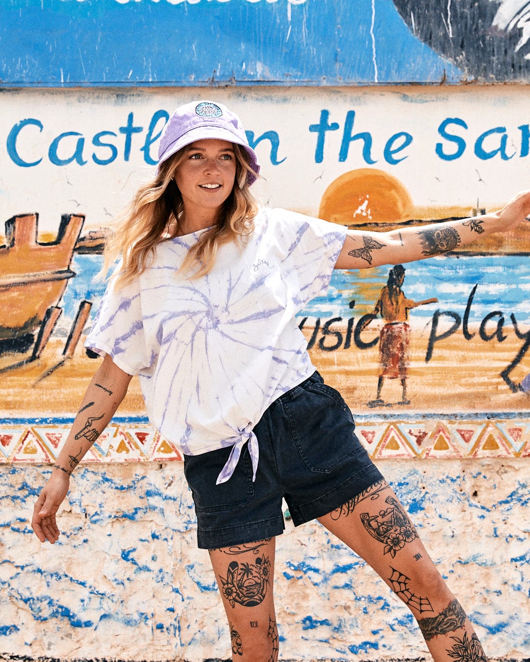 A young woman in a Swirl - Womens Short Sleeve T-Shirt - Tie Dye White/Purple by Saltrock and denim shorts smiles in front of a colorful mural titled "castle in the sand." She wears a cap and is heavily tattooed.