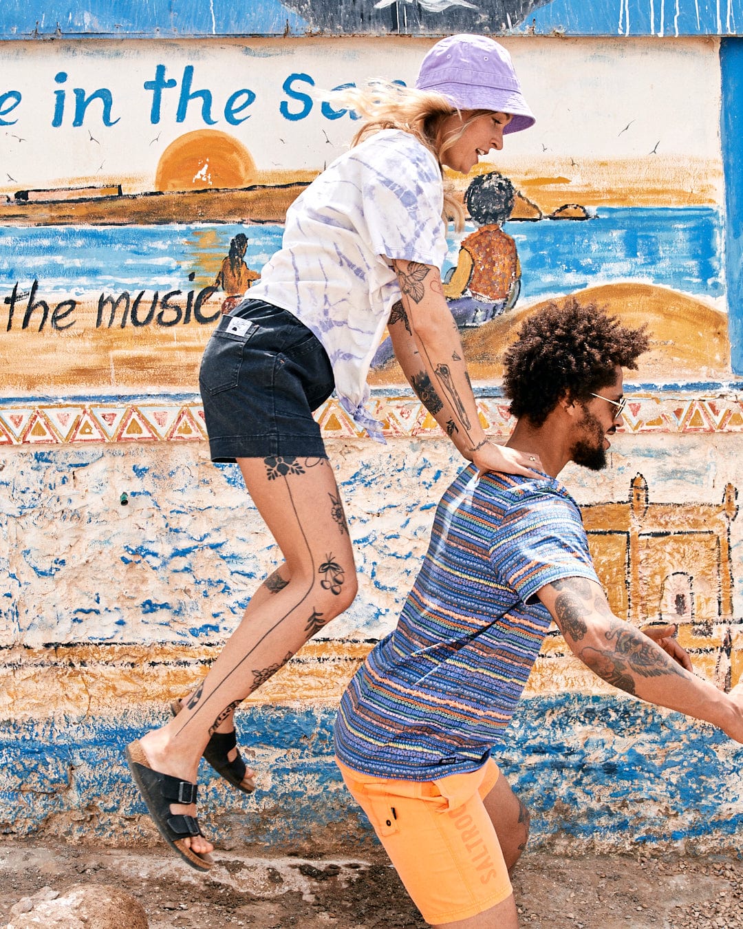 A young couple wearing Saltrock's Swirl - Womens Short Sleeve T-Shirt in Tie Dye White/Purple playfully dances in front of a vibrant mural with the phrase 'wave in the sea' written above.