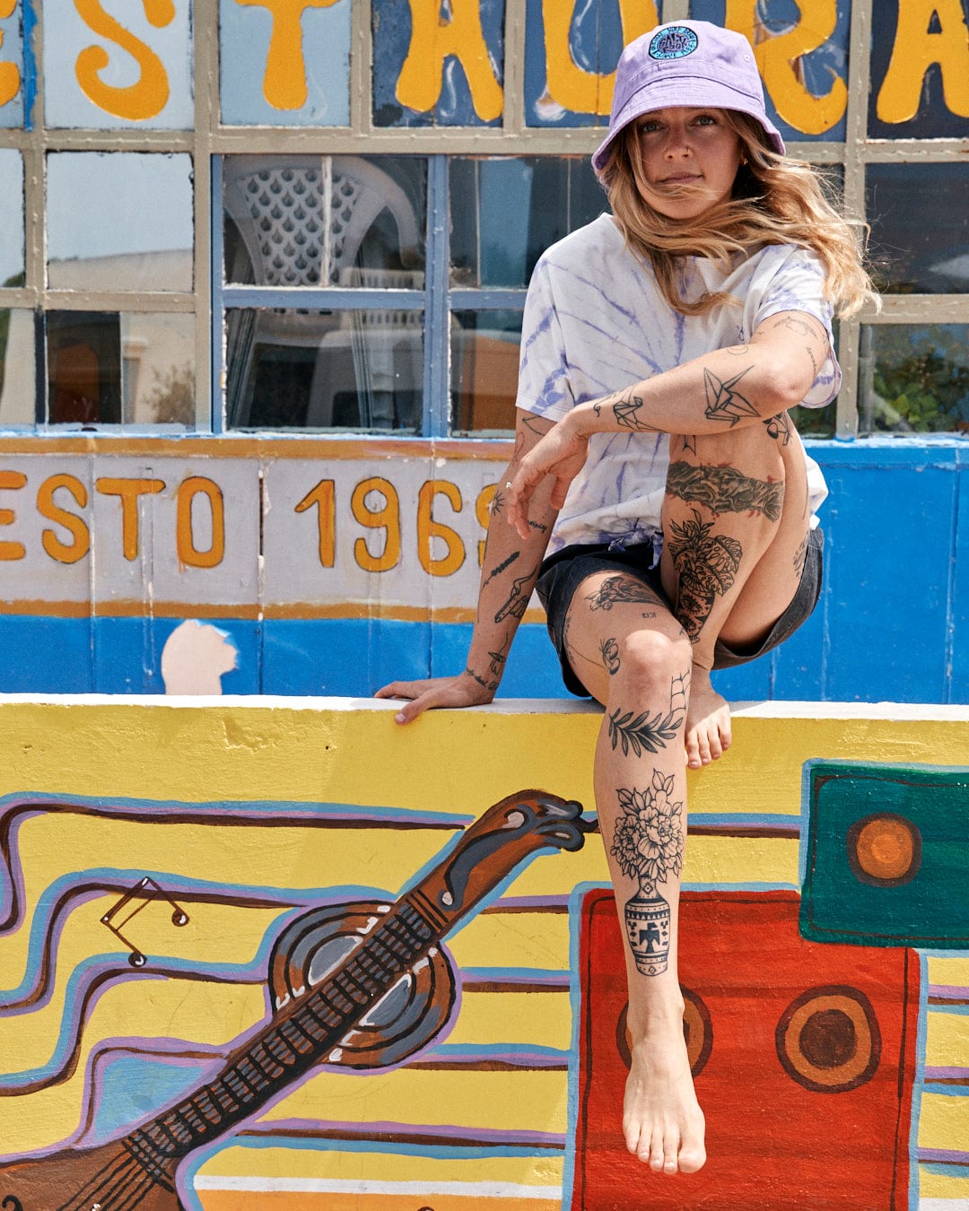 A woman with tattoos sits on a colorful painted ledge in front of a window with the text "esto 1966," wearing a lilac cap and a Saltrock Swirl - Women's Short Sleeve T-Shirt - Tie Dye White/Purple.