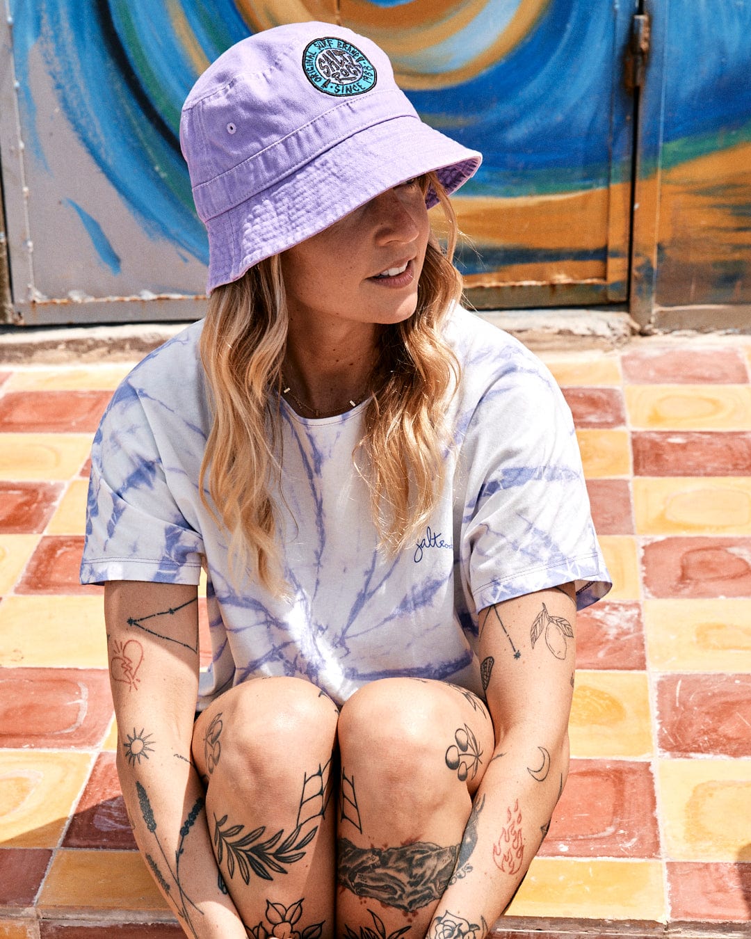 Woman in a purple bucket hat and blue Saltrock Swirl tie-dye short sleeve t-shirt sitting on a tiled step, looking to the side, with colorful graffiti in the background.