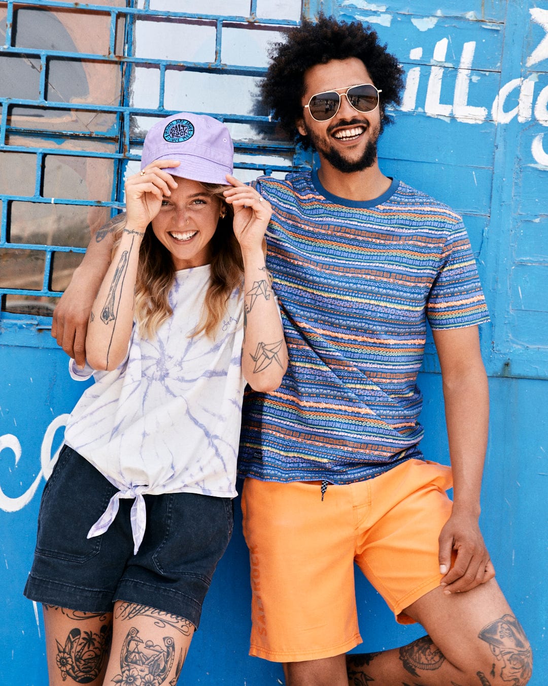 A joyful couple leaning against a blue wall; the woman adjusts her cap, and the man in a Saltrock Swirl Women's Short Sleeve T-Shirt in Tie Dye White/Purple wears sunglasses. Both are casually dressed and smiling.