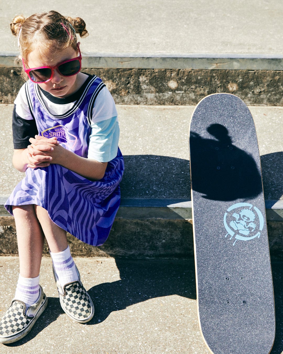 A child with short, styled hair sits on concrete steps, wearing a Saltrock Rezz - Kids Recycled Warp Vest Dress - Purple, red sunglasses, and checkered shoes. A skateboard with a cartoon graphic is placed beside them.