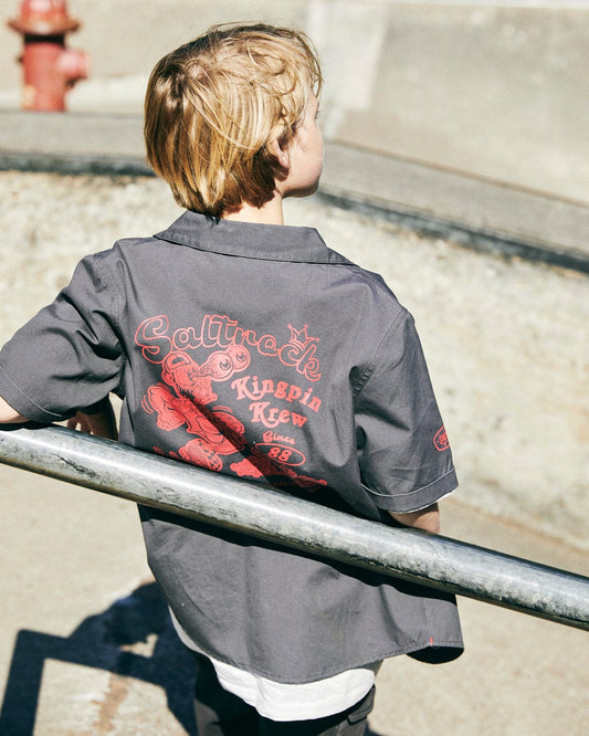 A child with blonde hair stands with his back to the camera, wearing a Kingpin Krew - Kids Short Sleeve Shirt - Dark Grey featuring a red Saltrock skateboard logo design that reads "Saltrock Kingpin.
