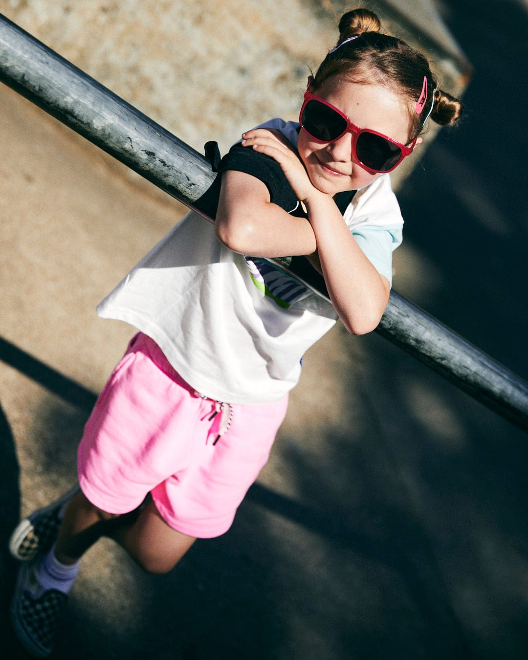 A child with double buns and red sunglasses leans on a metal railing, wearing a Saltrock Soda logo tee, pink shorts, and checkered slip-ons.