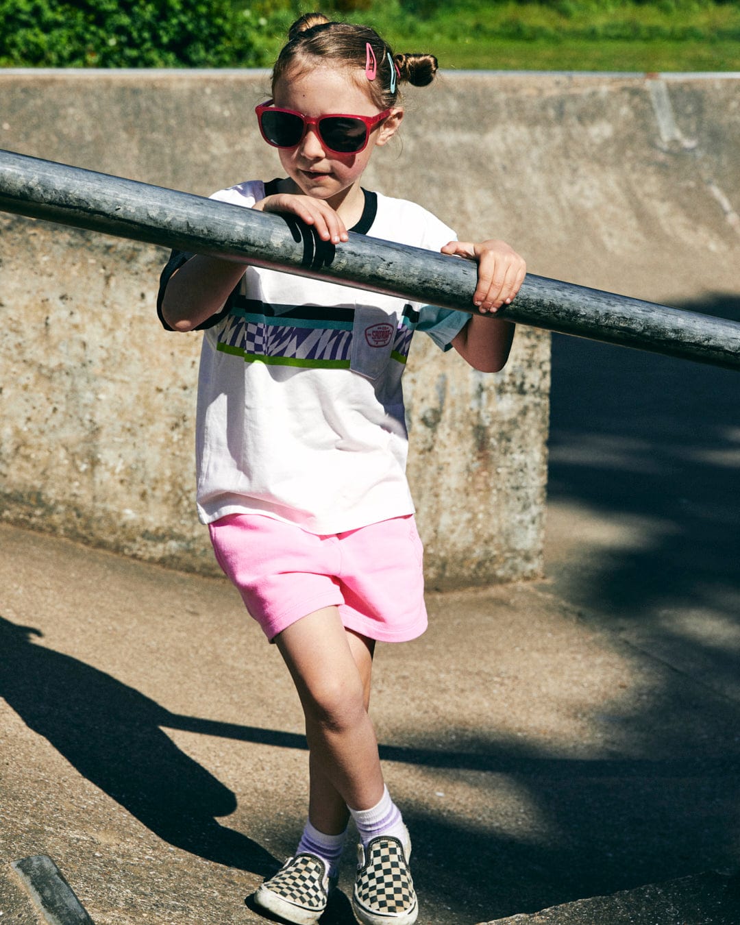 A child in a Saltrock Soda Recycled Kids Short Sleeve T-Shirt, sporting sunglasses and pink shorts, stands confidently on a skateboard ramp. Their checkered slip-on shoes grip the concrete as they hold a metal rail. The Saltrock logo gleams under the sunlight, casting playful shadows below.