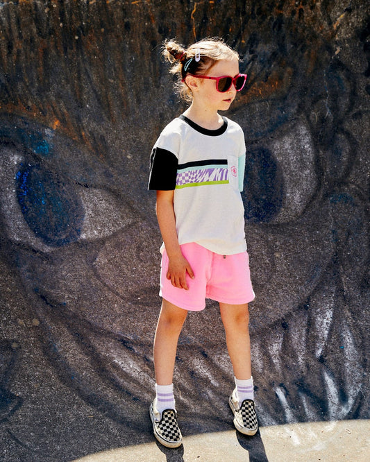 A child in sunglasses stands against a vibrant mural, wearing a colorblocked Soda Recycled Kids Short Sleeve T-Shirt by Saltrock, paired with pink shorts and checkered shoes.