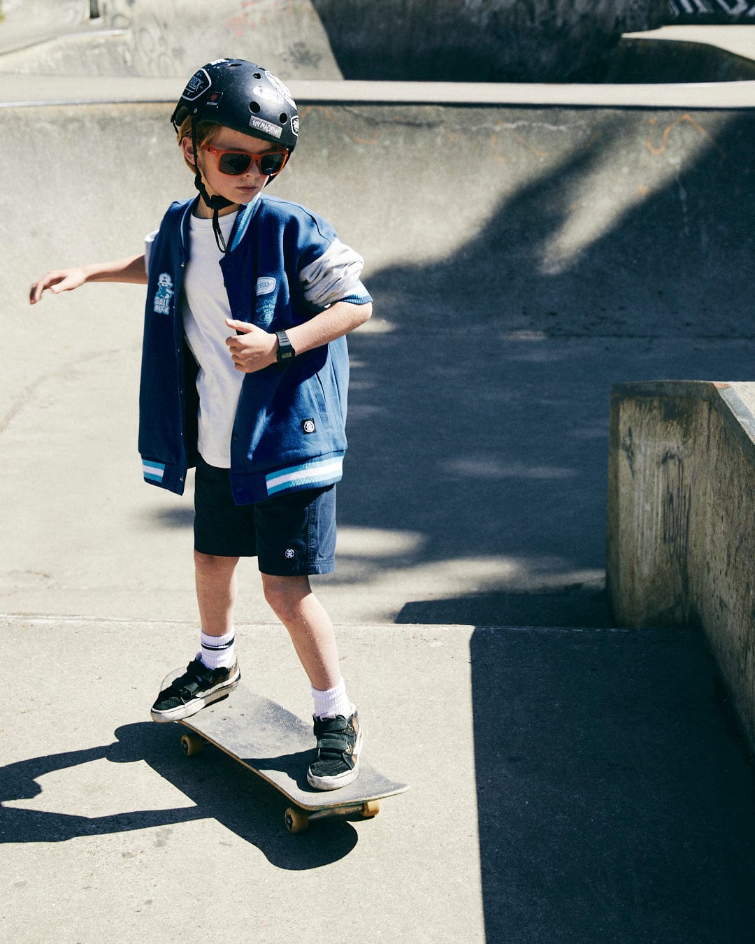 A young child wearing the Saltrock Krew Recycled Kids Varsity Bomber Jacket in blue, along with a helmet and sunglasses, skateboards in a concrete skate park.