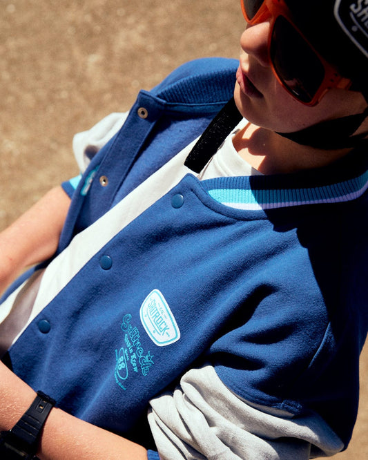 A child wears the Krew - Recycled Kids Varsity Bomber Jacket in blue by Saltrock, paired with trendy sunglasses and a black beanie featuring text.
