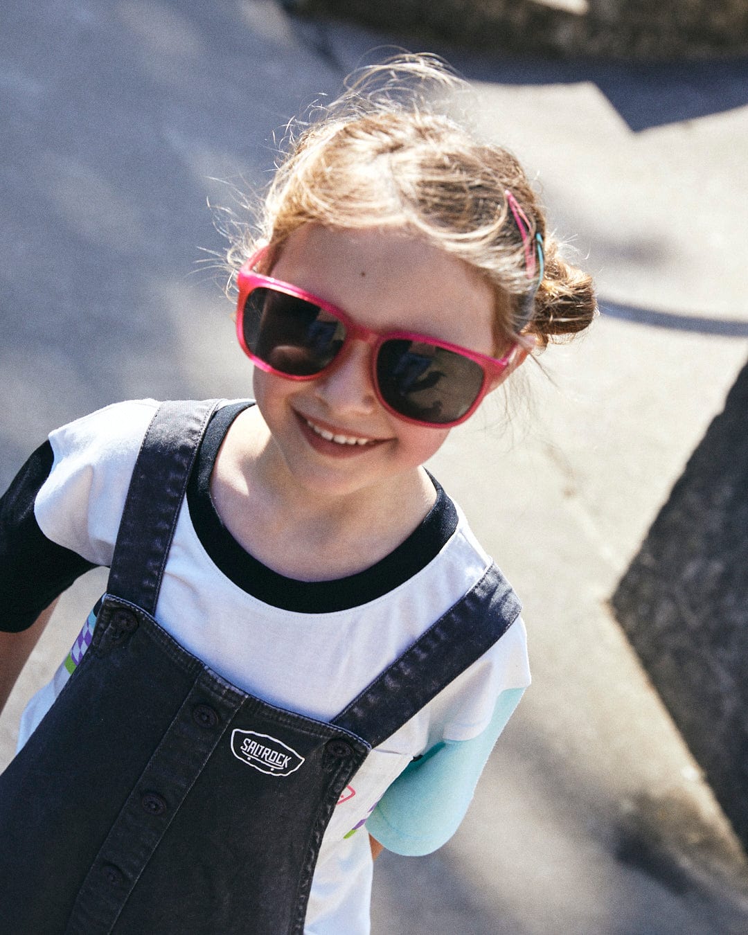 A young child in a white Soda Recycled Kids Short Sleeve T-Shirt by Saltrock, red sunglasses, and denim overalls smiles under the sunlight outdoors.
