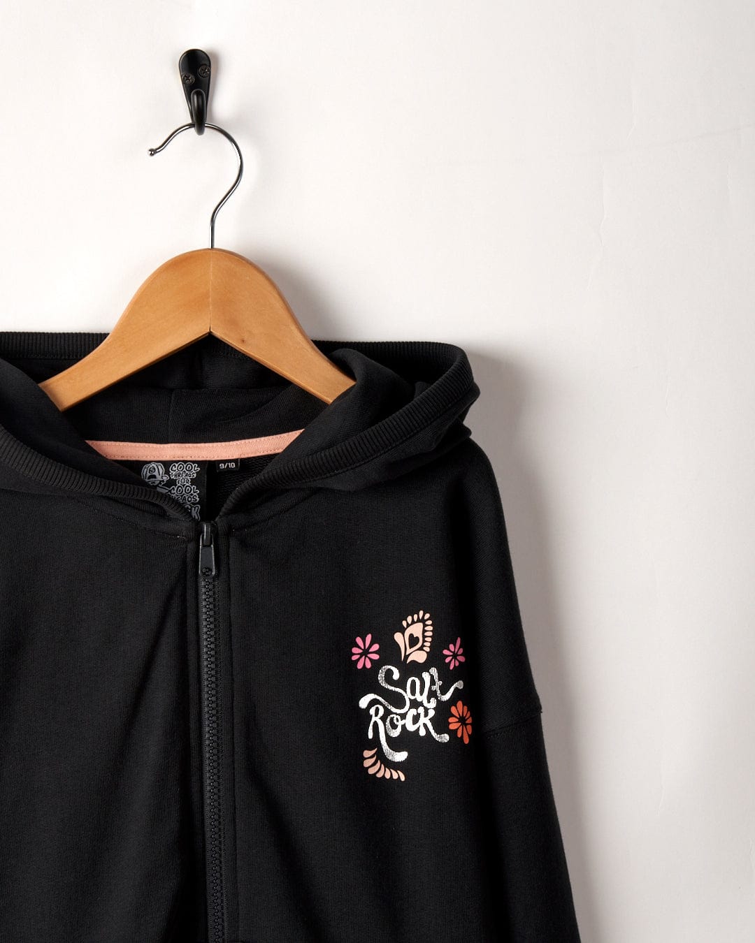 A Tahiti Van - Kids Zip Hoodie - Washed Black with a colorful skull and floral design, labeled "Saltrock branding," hangs on a wooden hanger against a white wall.