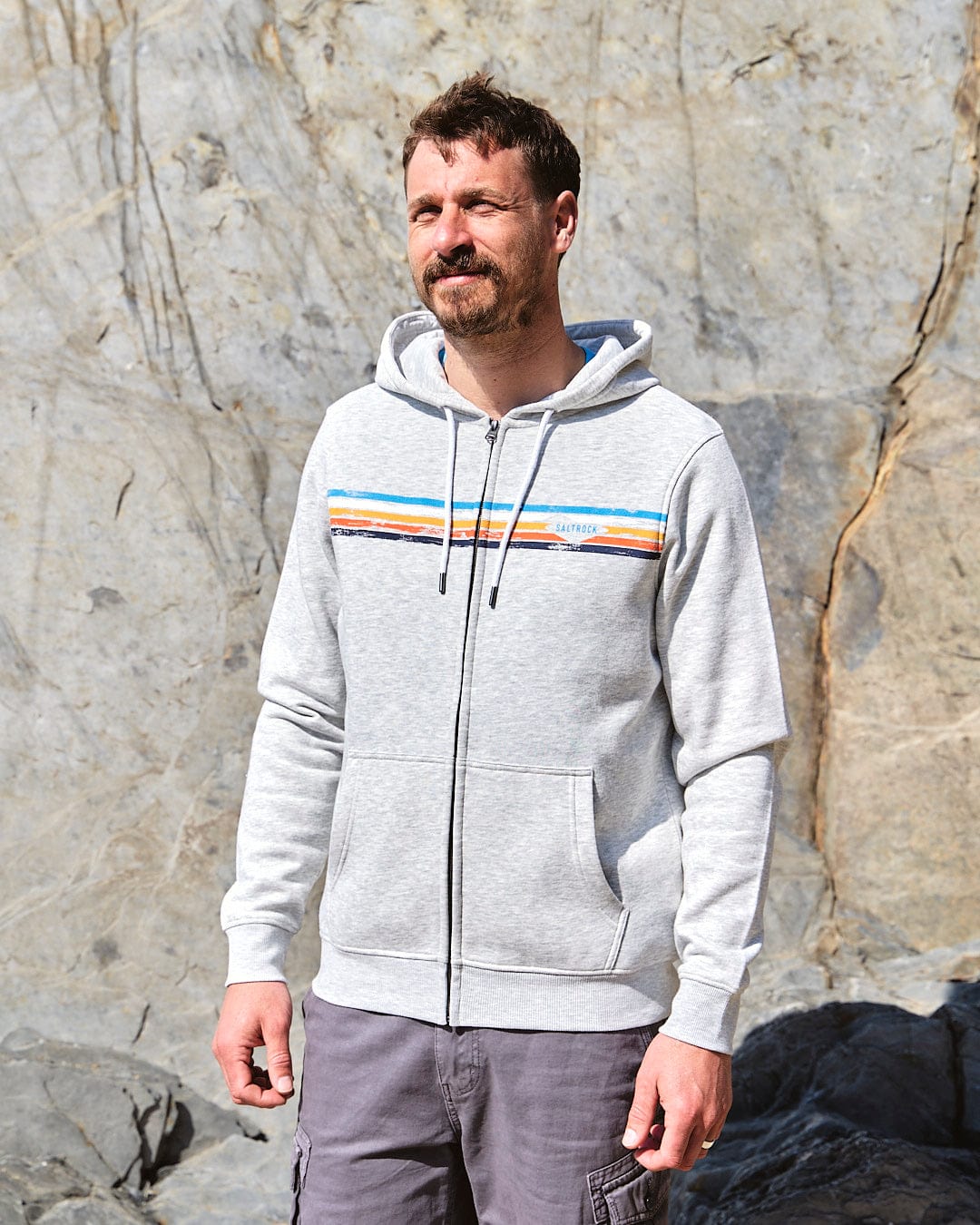 A man in a Saltrock Taped Stripe - Mens Zip Hoodie - Grey standing next to a rock.