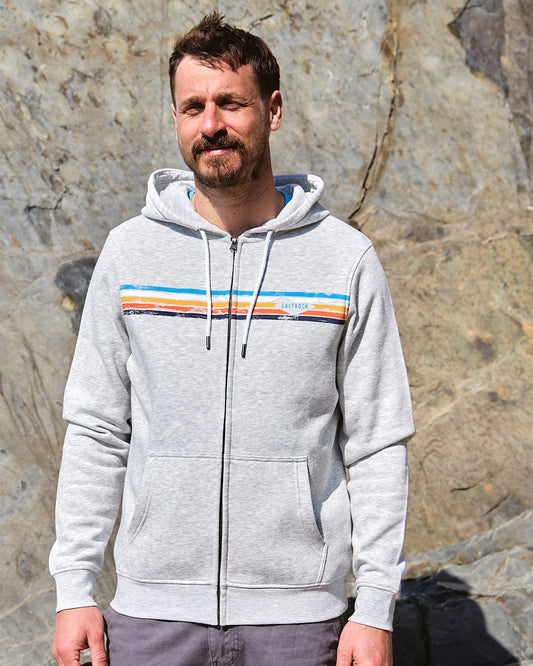 A man wearing a Saltrock Taped Stripe - Mens Zip Hoodie - Grey in front of a rock.