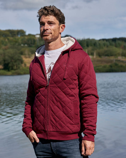 A man wearing a Tarka - Mens Quilted Borg Lined Hoody - Red, branded with Saltrock, standing next to a lake.