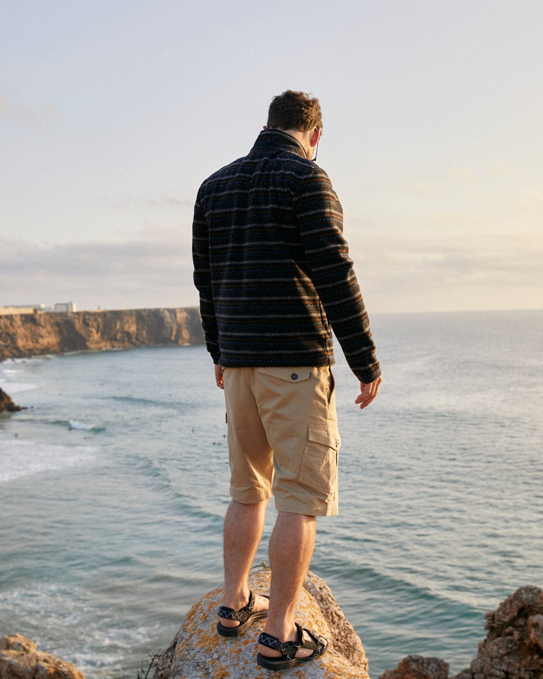 A man standing on a rock overlooking a body of water wearing a Saltrock Theo Stripe - Mens Fleece - Dark Grey Stripe.
