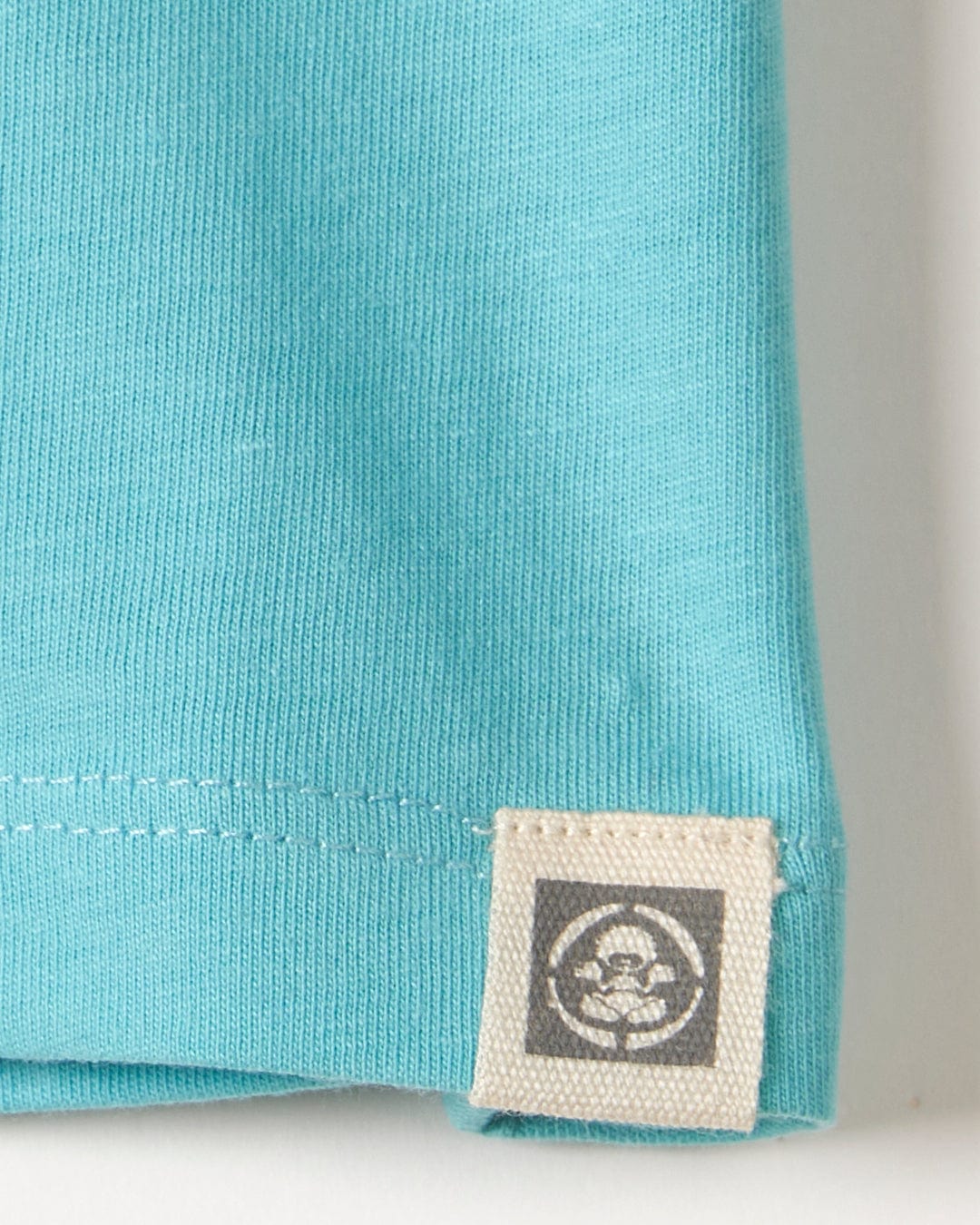 Close-up of a teal Tok Corp Recycled Kids Short Sleeve T-Shirt fabric with a small white label featuring a circular Saltrock branding logo, attached to the hem.