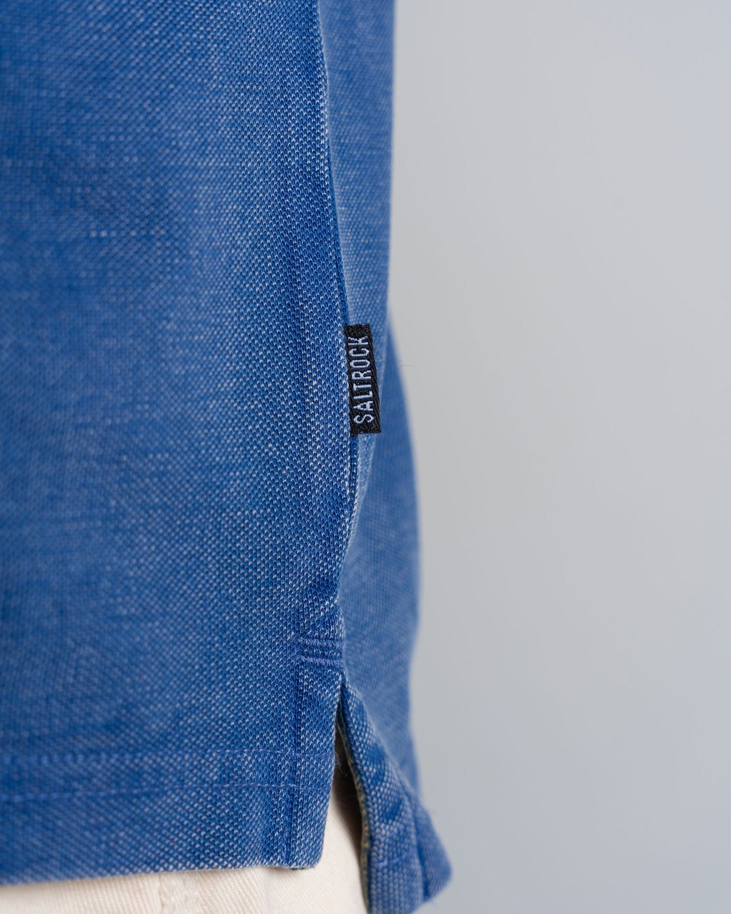 Close-up of a blue textured 100% cotton garment, featuring a side seam with a small black Saltrock logo tag; the bottom edge of the Seb - Mens Short Sleeve Polo - Blue has a slight slit above beige fabric.