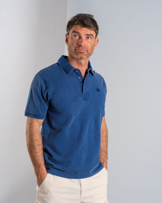 A man stands with hands in pockets, looking slightly to the side, wearing a Saltrock "Seb" men's short sleeve blue polo and beige shorts against a neutral background.